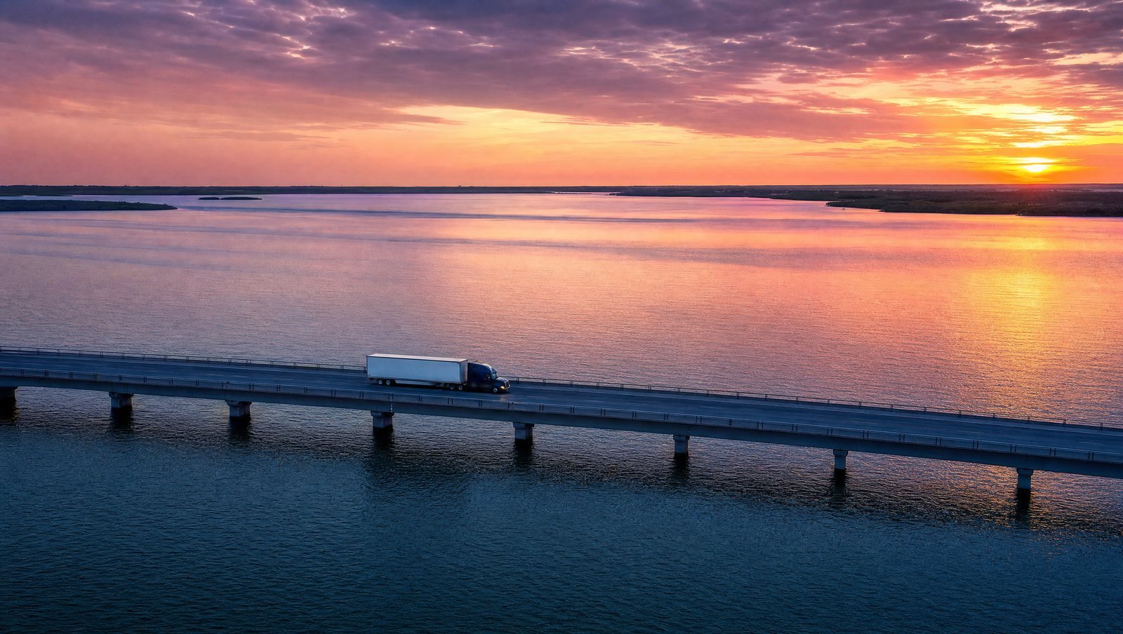 Semi truck hauling a trailer across a long bridge over open water at sunset